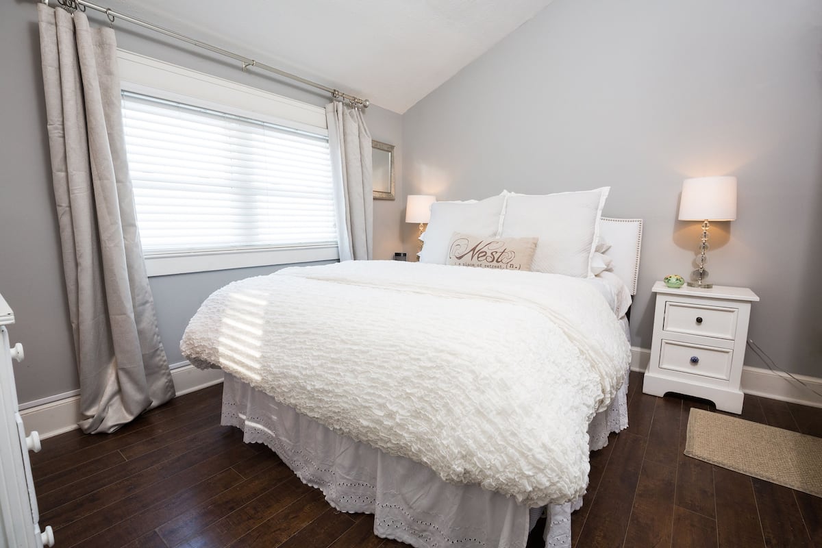 A bedroom with a large bed, white bedding, a nightstand with a lamp, and a window with curtains.