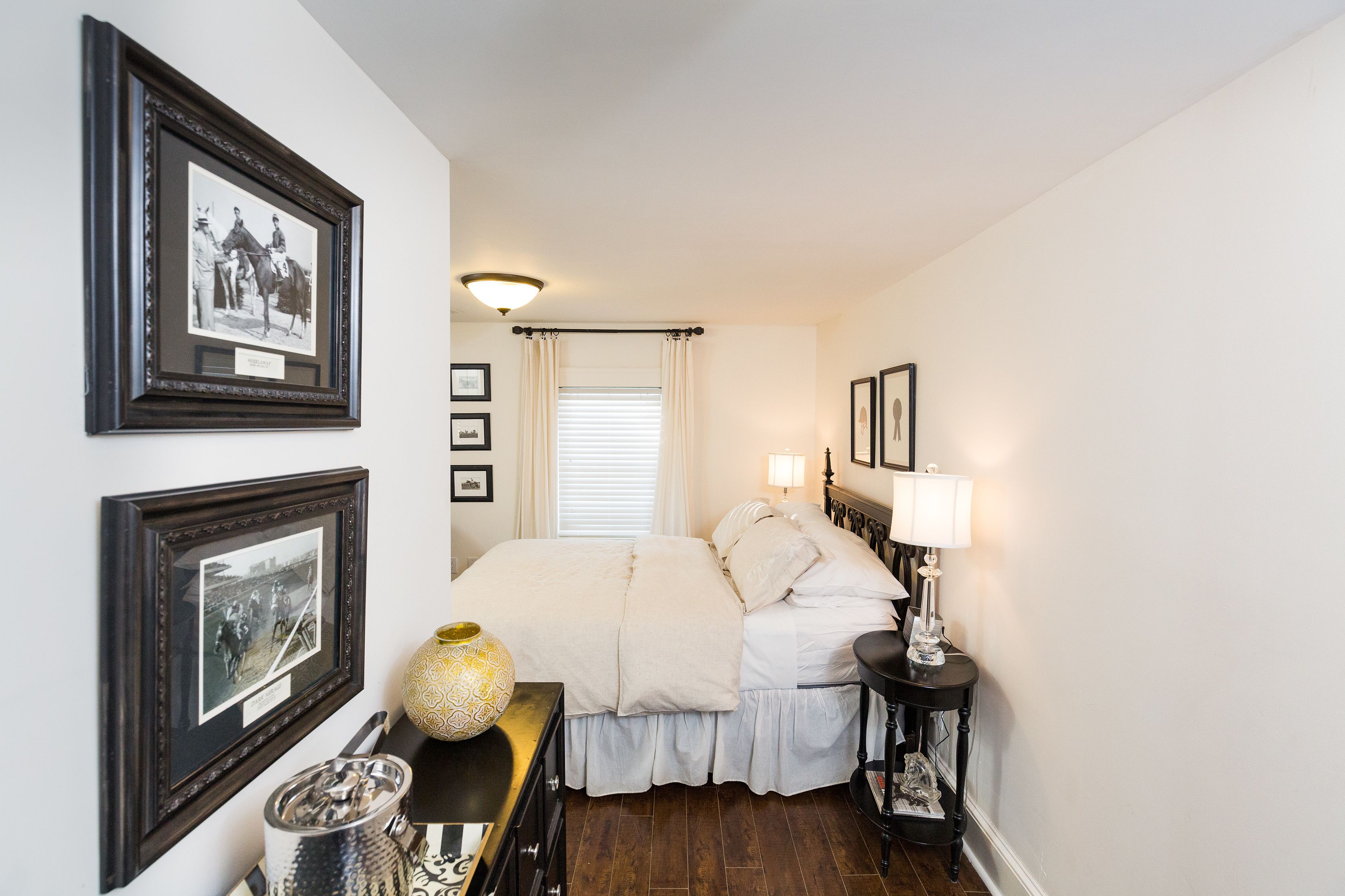 A bedroom with a large bed, framed pictures on the walls, a bedside table with a lamp, and a window with curtains.
