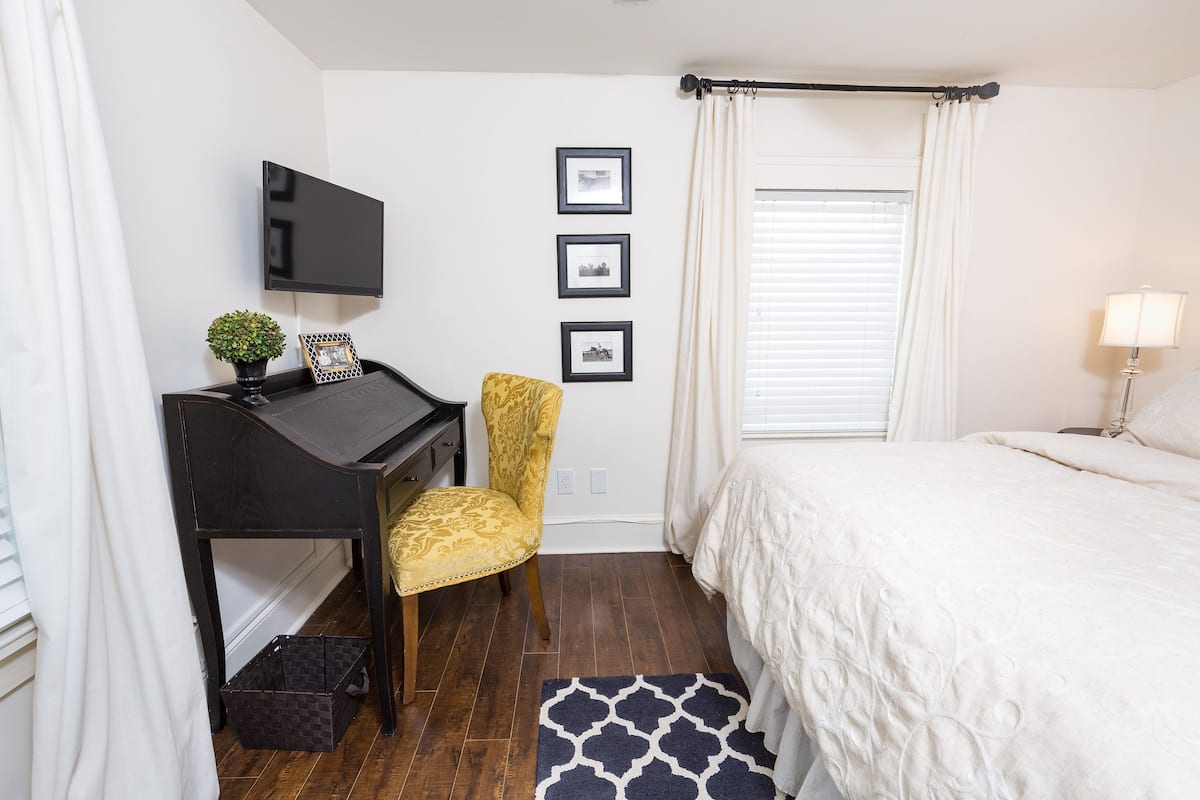 A bedroom with a bed, a desk, a chair, a TV, and a window with curtains.