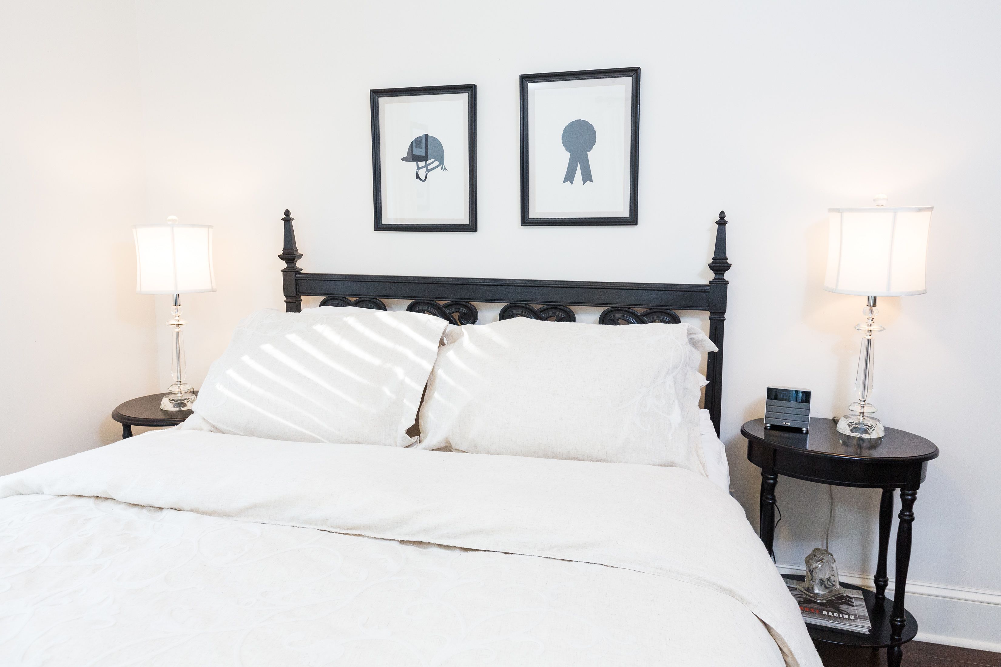 Una cama bien hecha con sábanas blancas, dos mesitas de noche con lámparas, y dos cuadros en la pared.