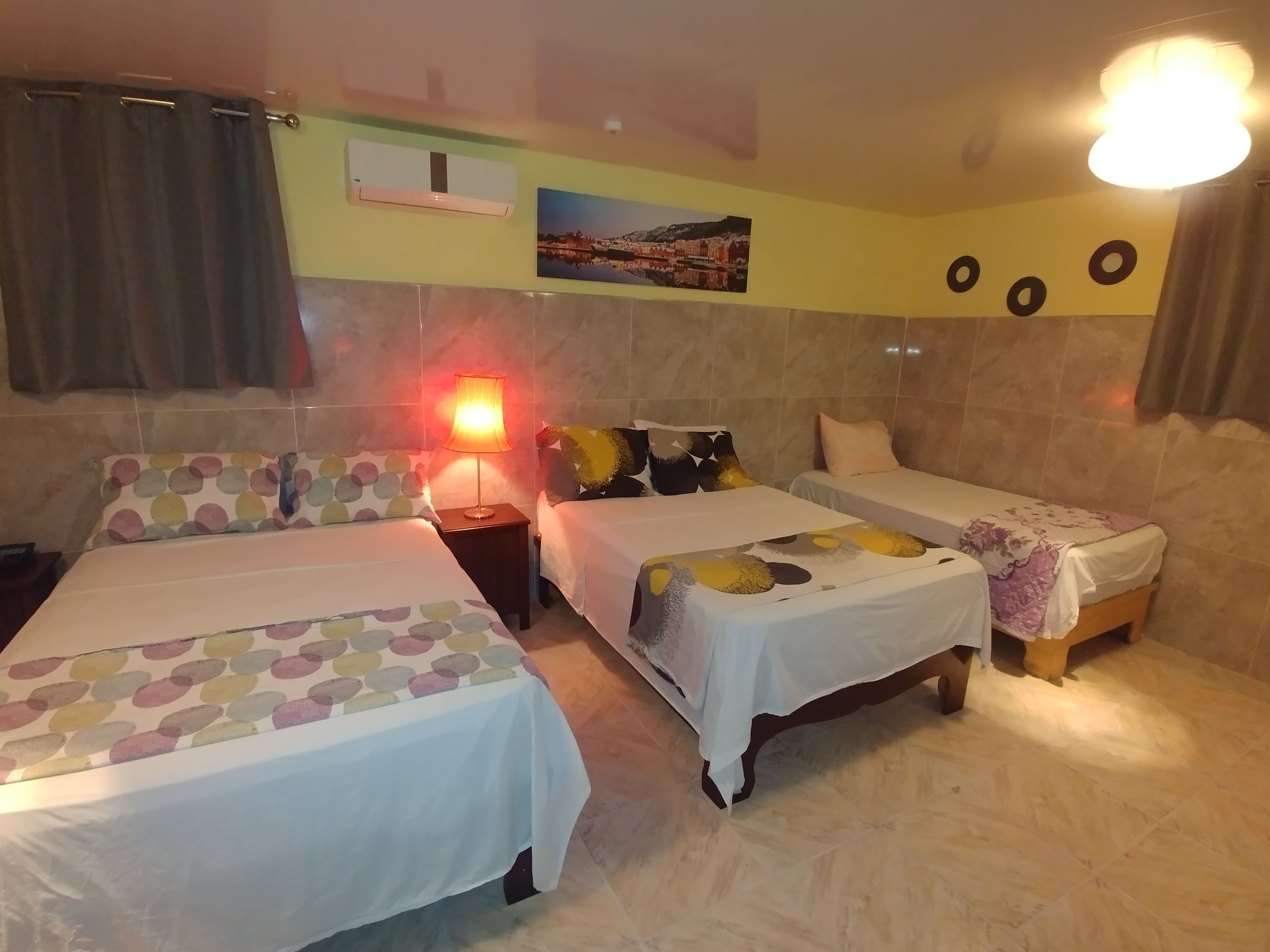 A hotel room with two beds, a nightstand with a lamp, a wall-mounted air conditioner, and a picture of a cityscape.