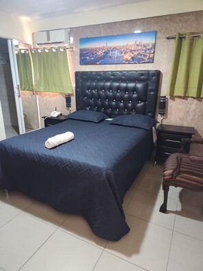 A bedroom with a large bed, a headboard, a nightstand, a chair, a painting, and curtains. - Hotel Riparbella (Santo Domingo)