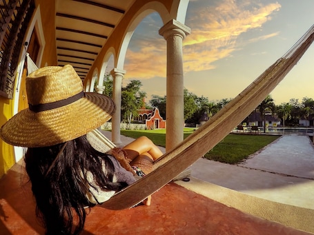 A person relaxing in a hammock under a covered porch with a view of a building and greenery.