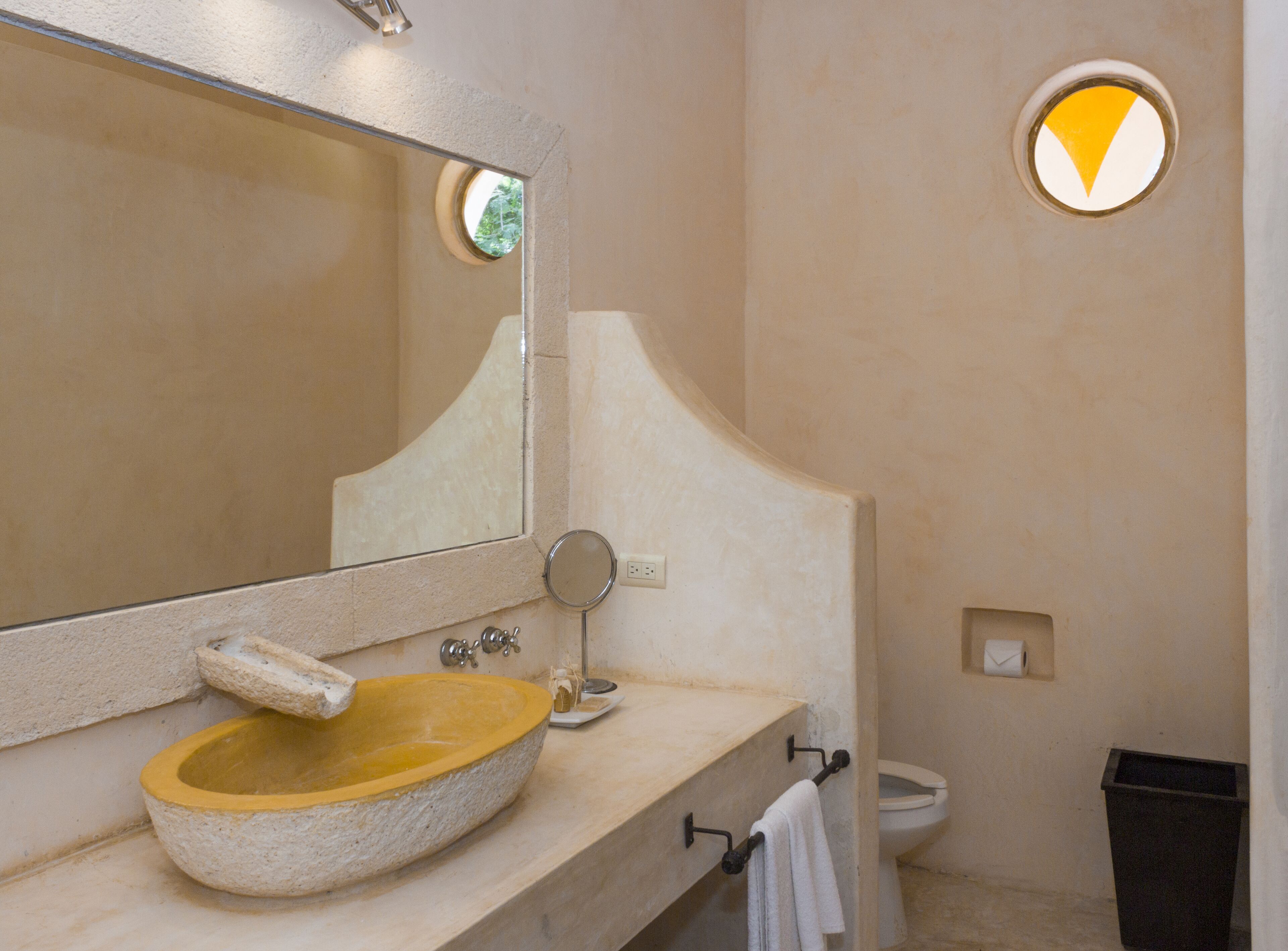 Un baño con un lavabo de piedra, un espejo grande, un espejo redondo pequeño, un perchero y un inodoro.