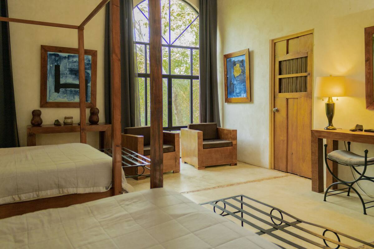A room with a four-poster bed, a desk with a lamp, two chairs, and artwork on the walls.