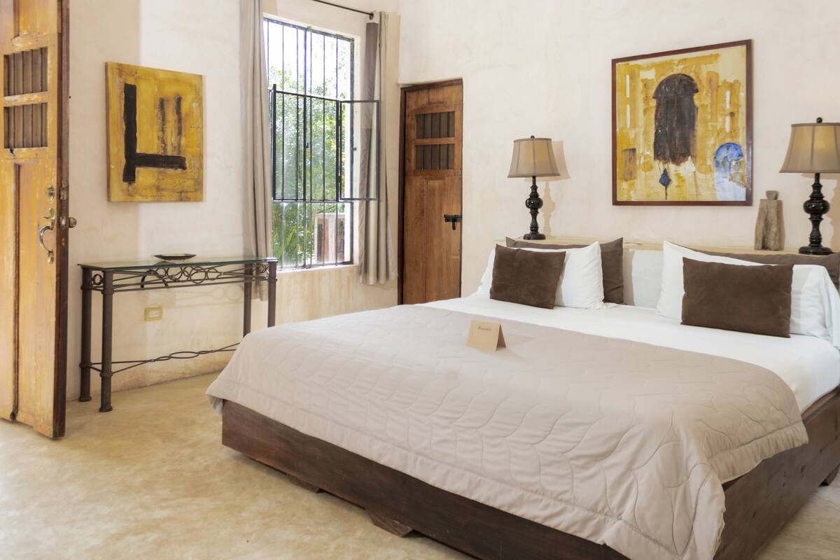 A bedroom with a large bed, a bedside table, two lamps, a window with bars, and two paintings on the wall.