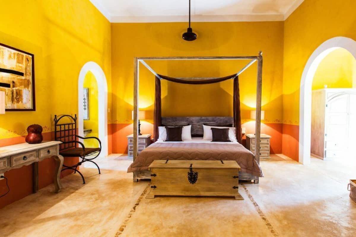 A room with a four-poster bed, two bedside tables, a desk with a chair, and a large window.