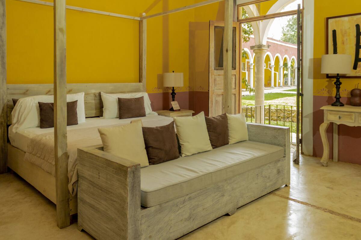 A room with a four-poster bed, a wooden bench, and a view of an outdoor courtyard with arches.