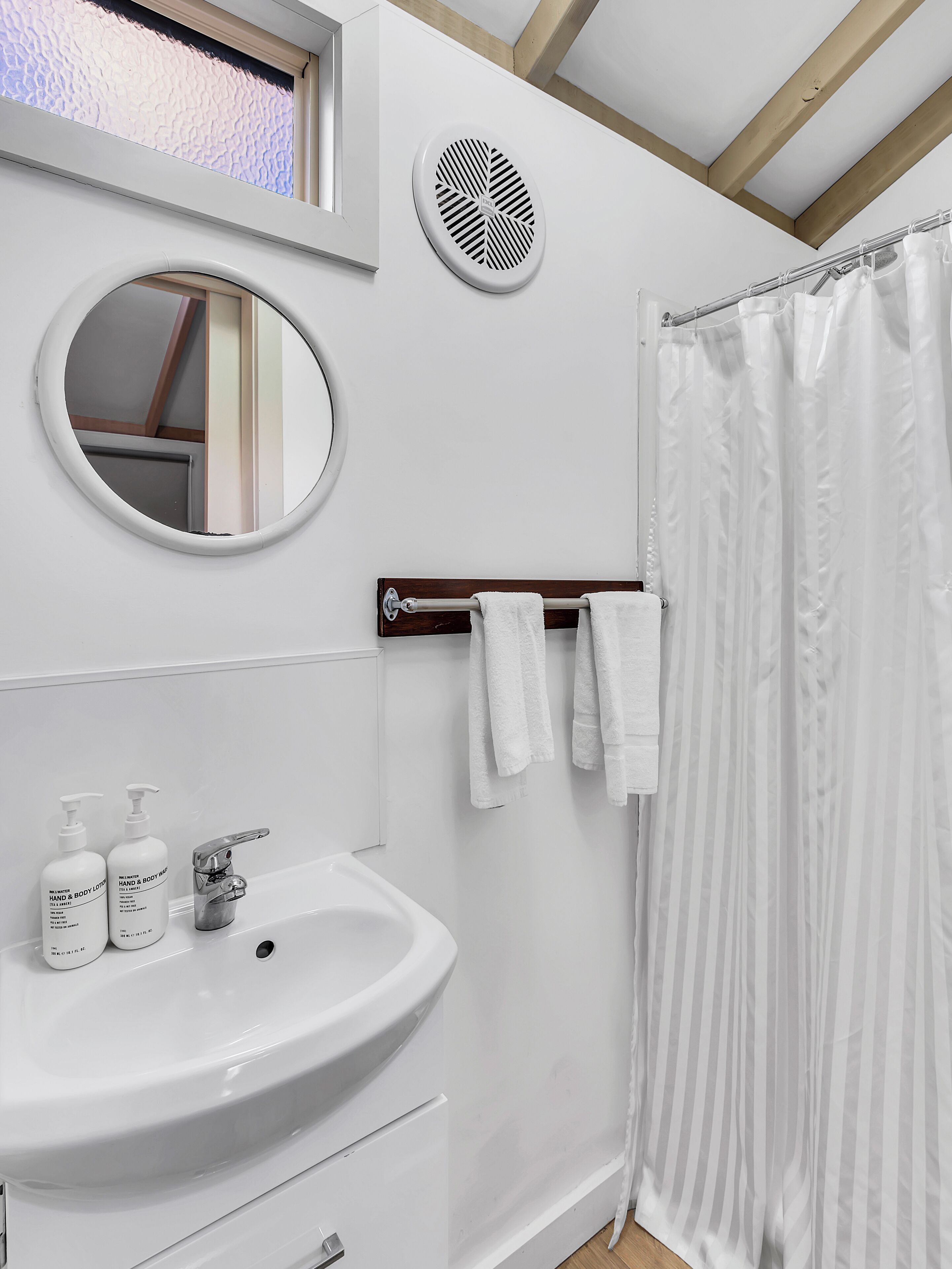 Un baño con un lavabo blanco, dos botellas de jabón de manos y cuerpo, un espejo, un toallero con toallas, una cortina de ducha y un respiradero circular en la pared.