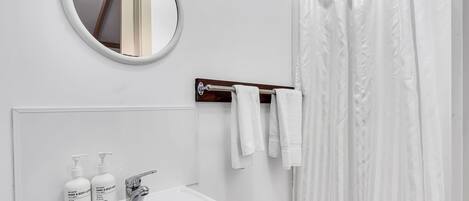 Un baño con un lavamanos blanco, dos botellas de crema para manos y cuerpo, un espejo y una cortina de ducha.