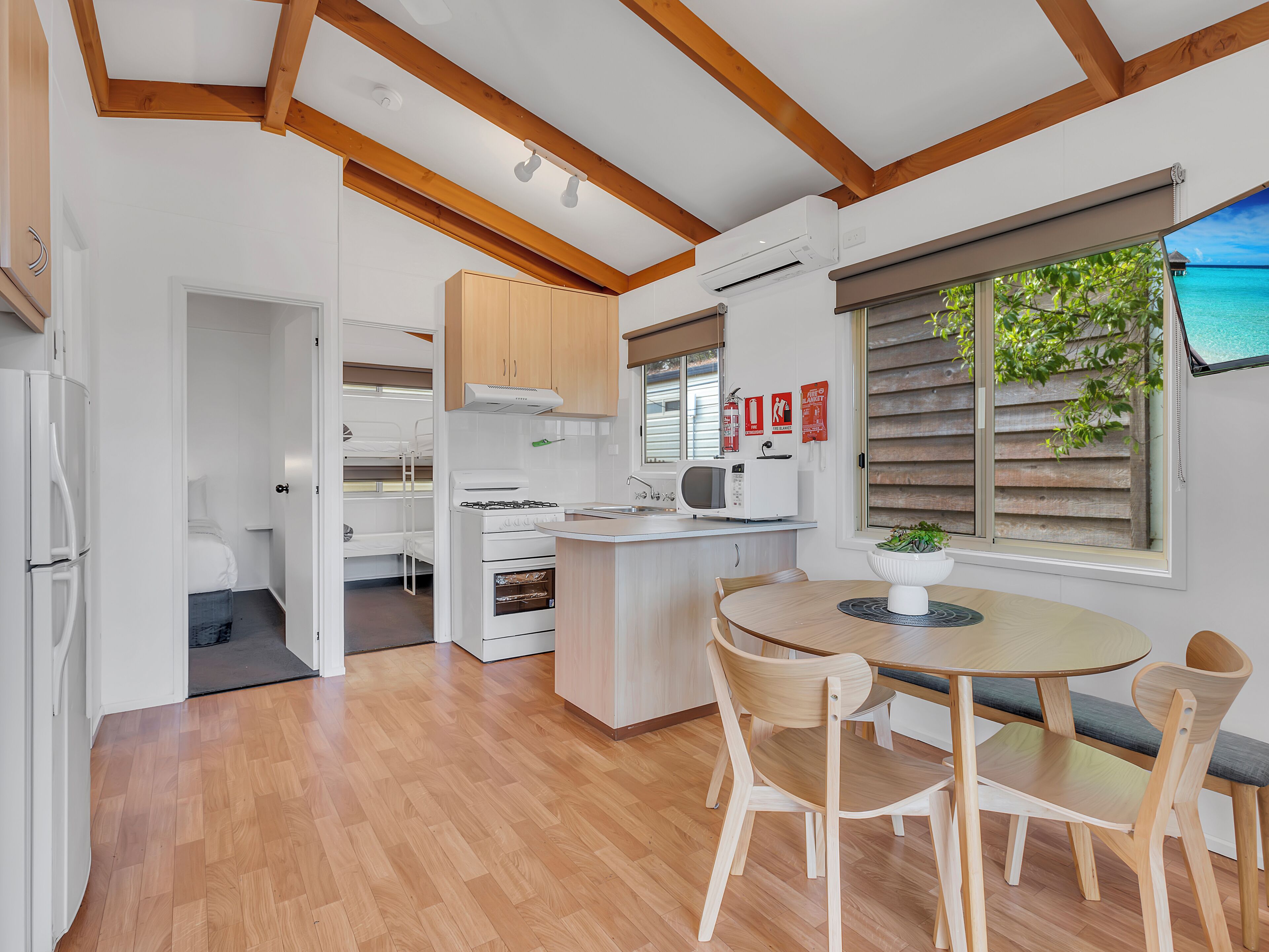 Une cuisine compacte avec des meubles en bois, une cuisinière et un four à micro-ondes. On y trouve un coin repas avec une table et des chaises. La pièce est dotée d’un plancher en bois et d’une fenêtre avec des stores.