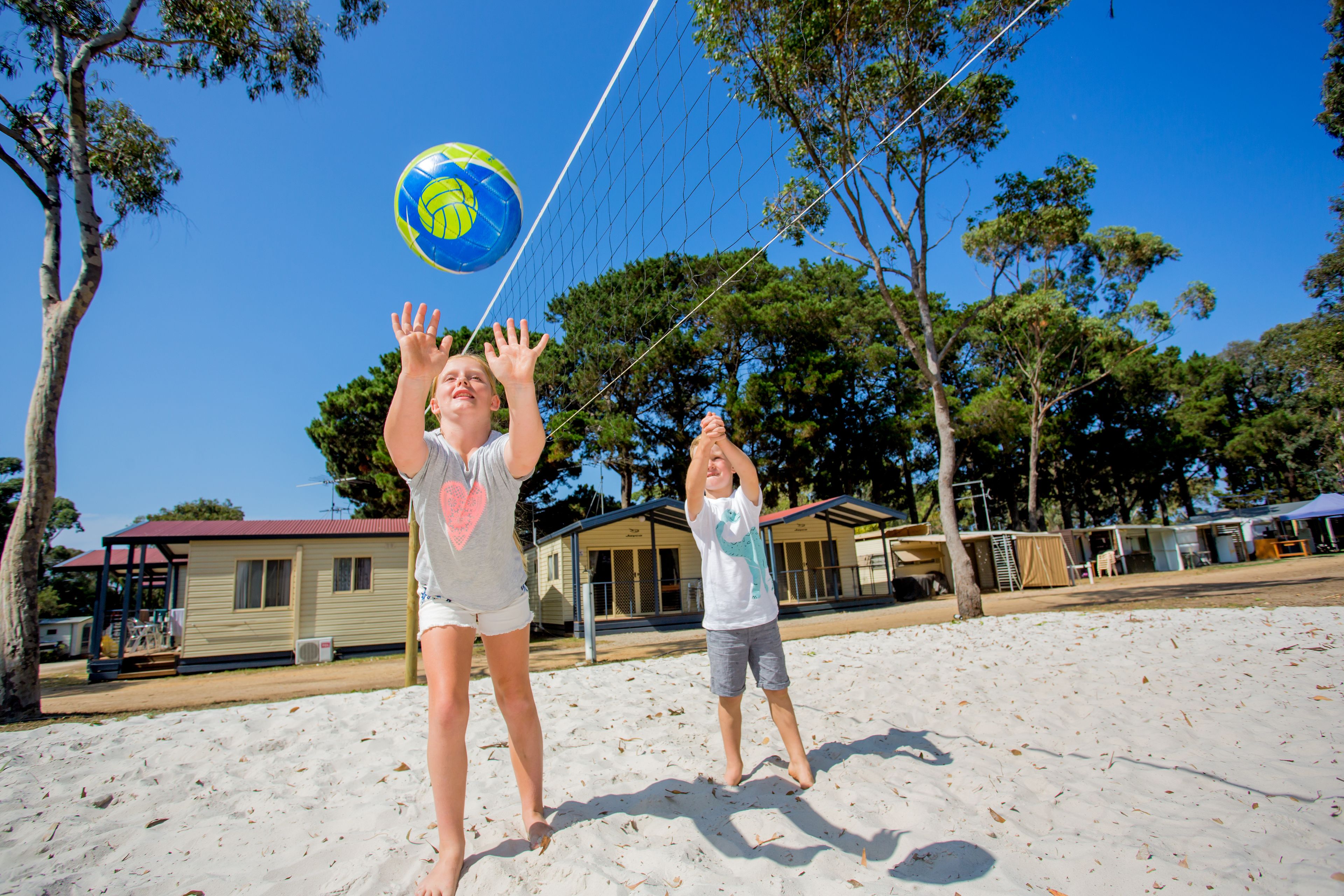 Dos personas jugando voleibol en una playa arenosa, con árboles y un edificio al fondo.