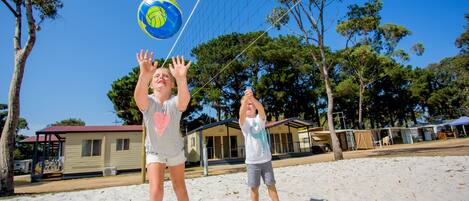 Dos personas jugando voleibol en una playa arenosa, con árboles y un edificio al fondo.