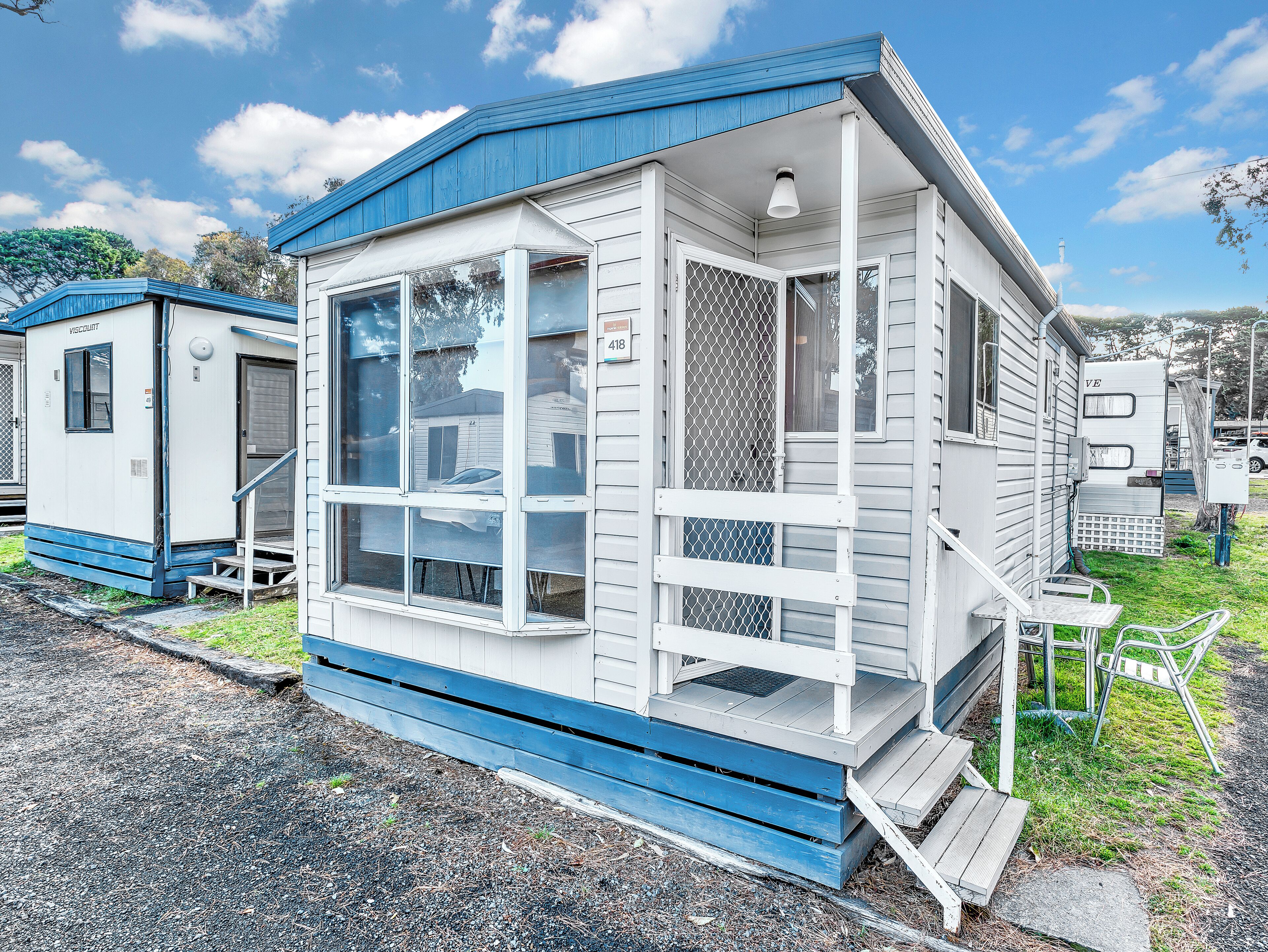 Una casa móvil pequeña de un solo piso con techo azul y revestimiento blanco, con ventanas grandes y un porche.