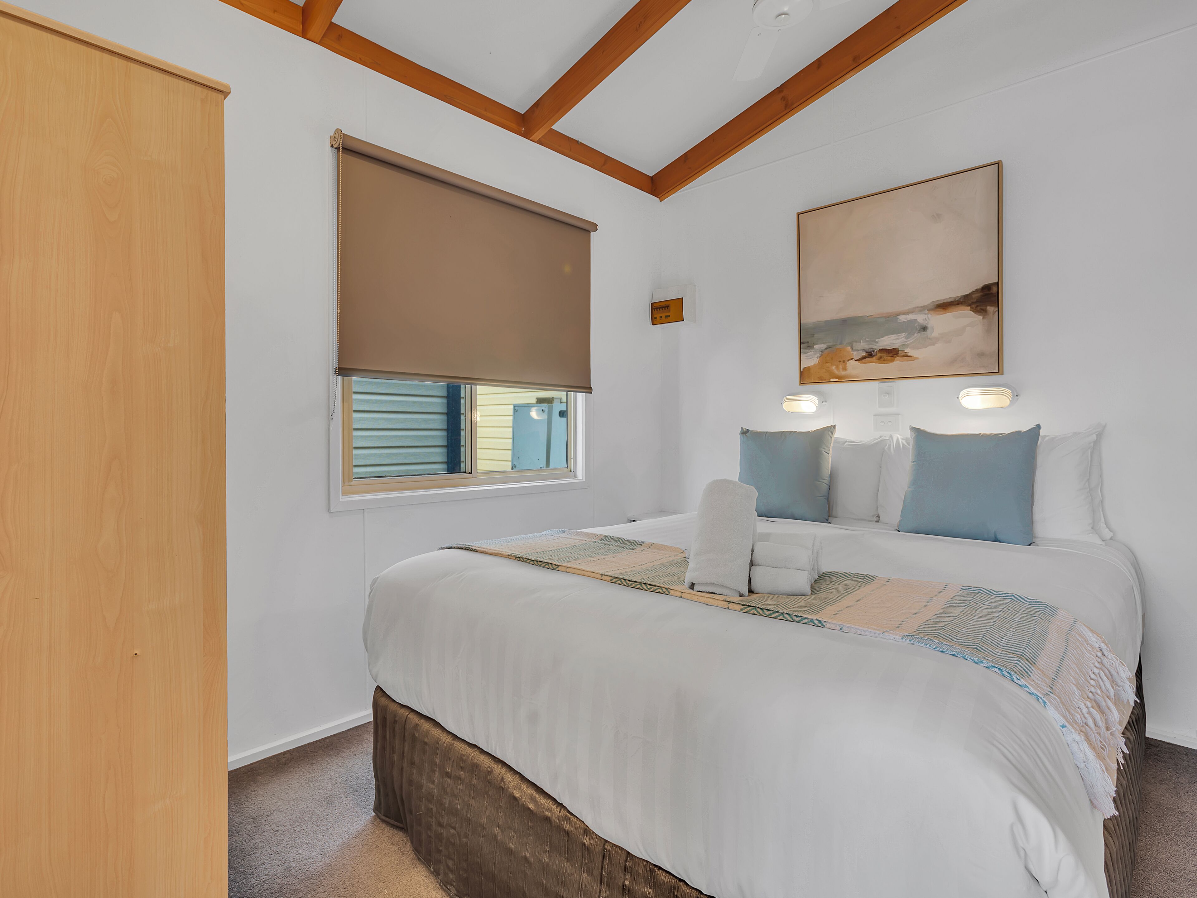 A bedroom with a bed, a window with blinds, a painting on the wall, and a wooden ceiling beam.