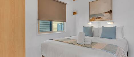A bedroom with a bed, a window with blinds, a painting on the wall, and a wooden ceiling beam.
