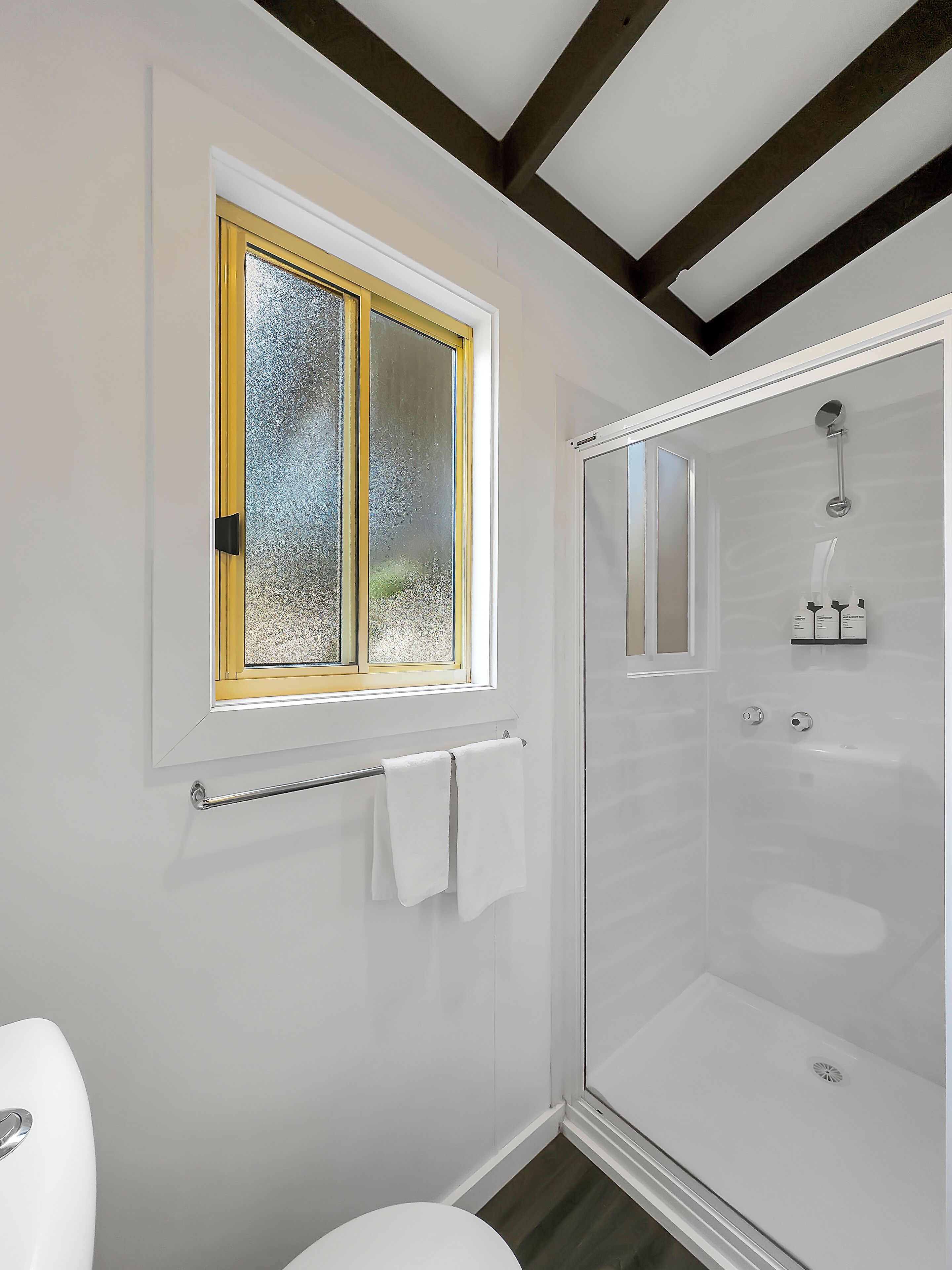 A bathroom with a glass shower enclosure, a toilet, and a window with a yellow frame.