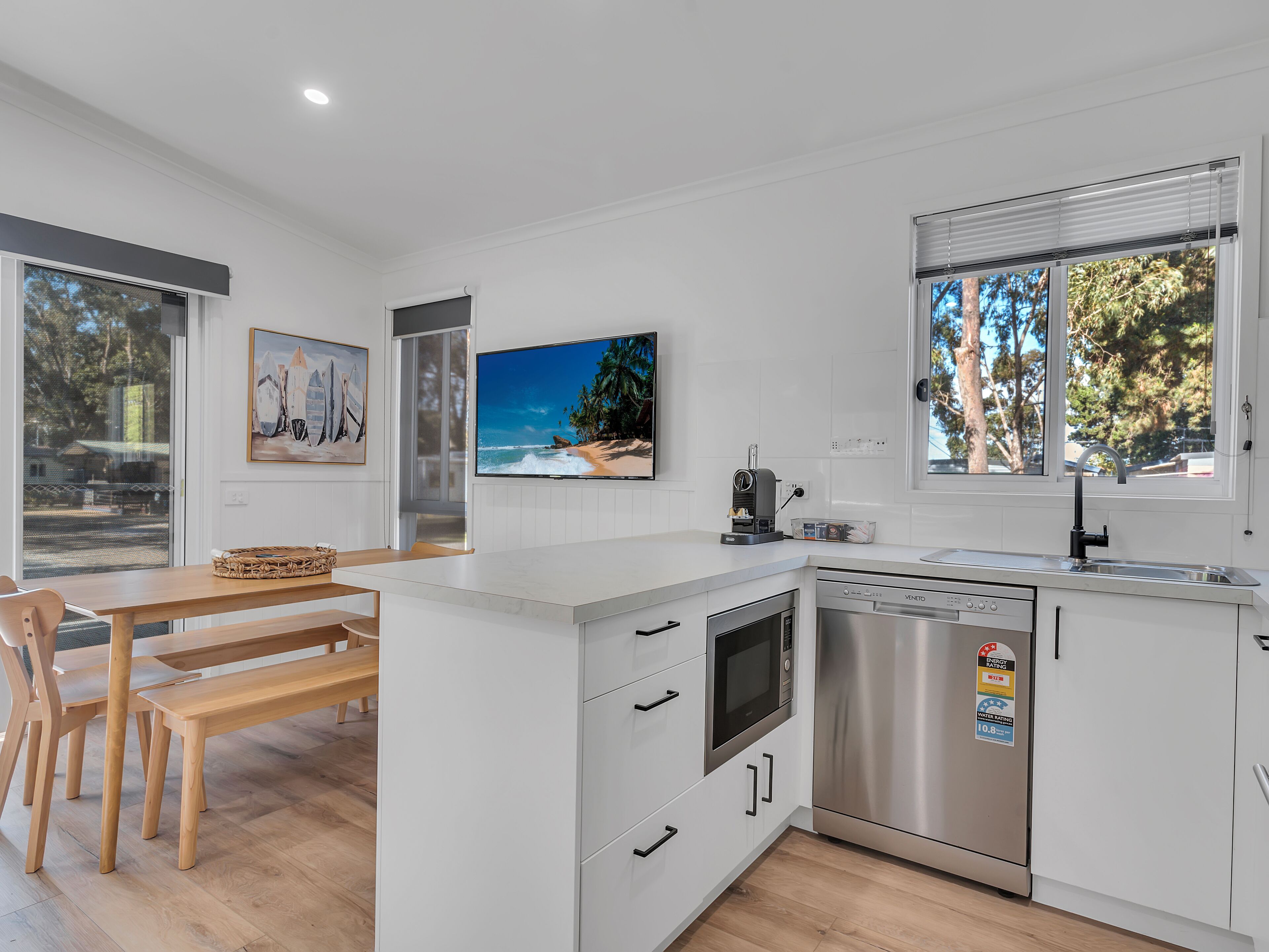 A modern kitchen with white cabinetry, a built-in oven, and a dishwasher. There is a dining area with a wooden table and chairs, and a large window with a view of trees.