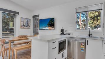 A modern kitchen with white cabinetry, a built-in oven, and a dishwasher. There is a dining area with a wooden table and chairs, and a large window with a view of trees.