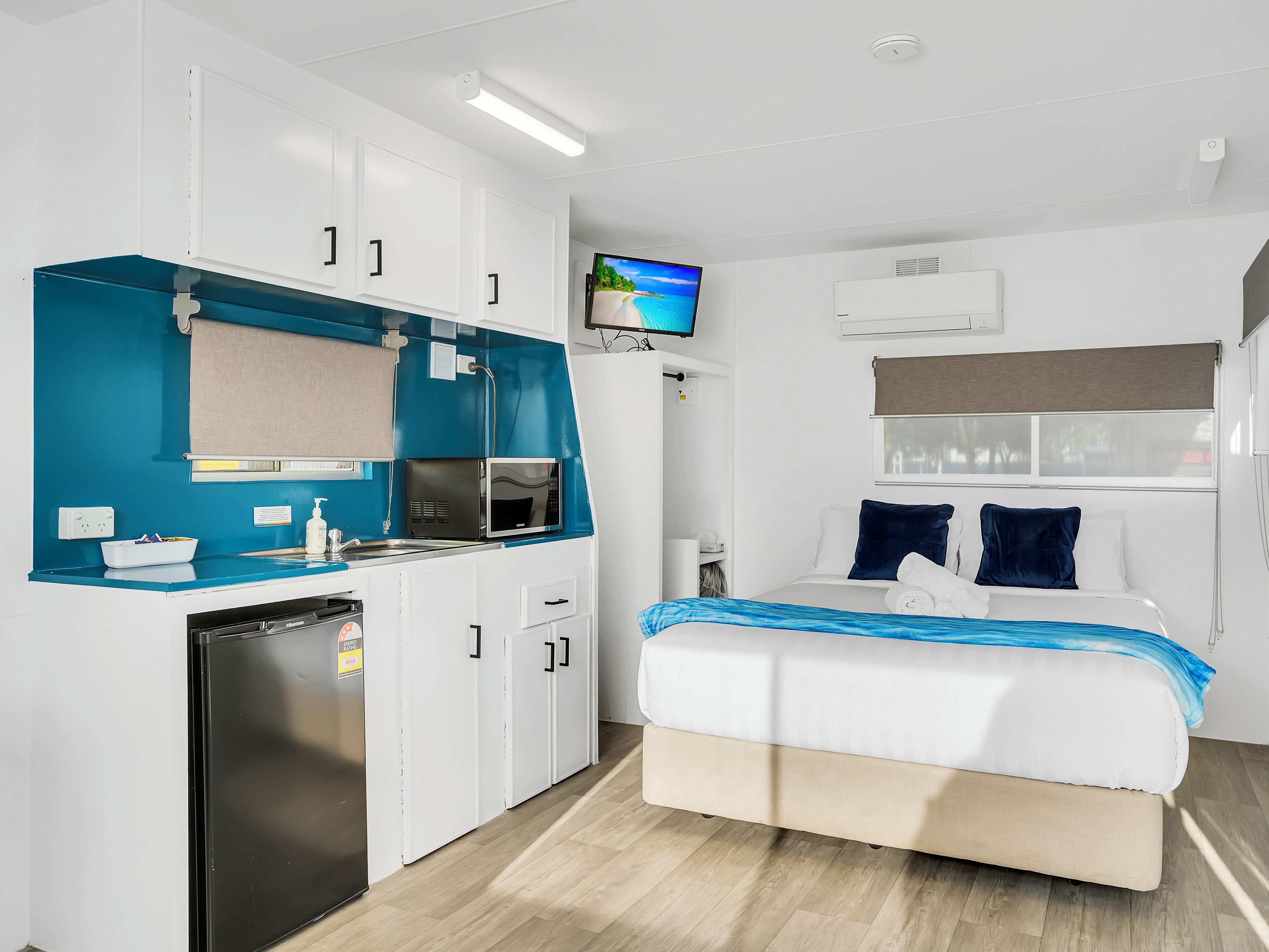 Une chambre d’hôtel compacte, dotée d’une kitchenette, d’un lit avec une literie bleue, d’une télévision et d’une fenêtre donnant sur un paysage agréable.