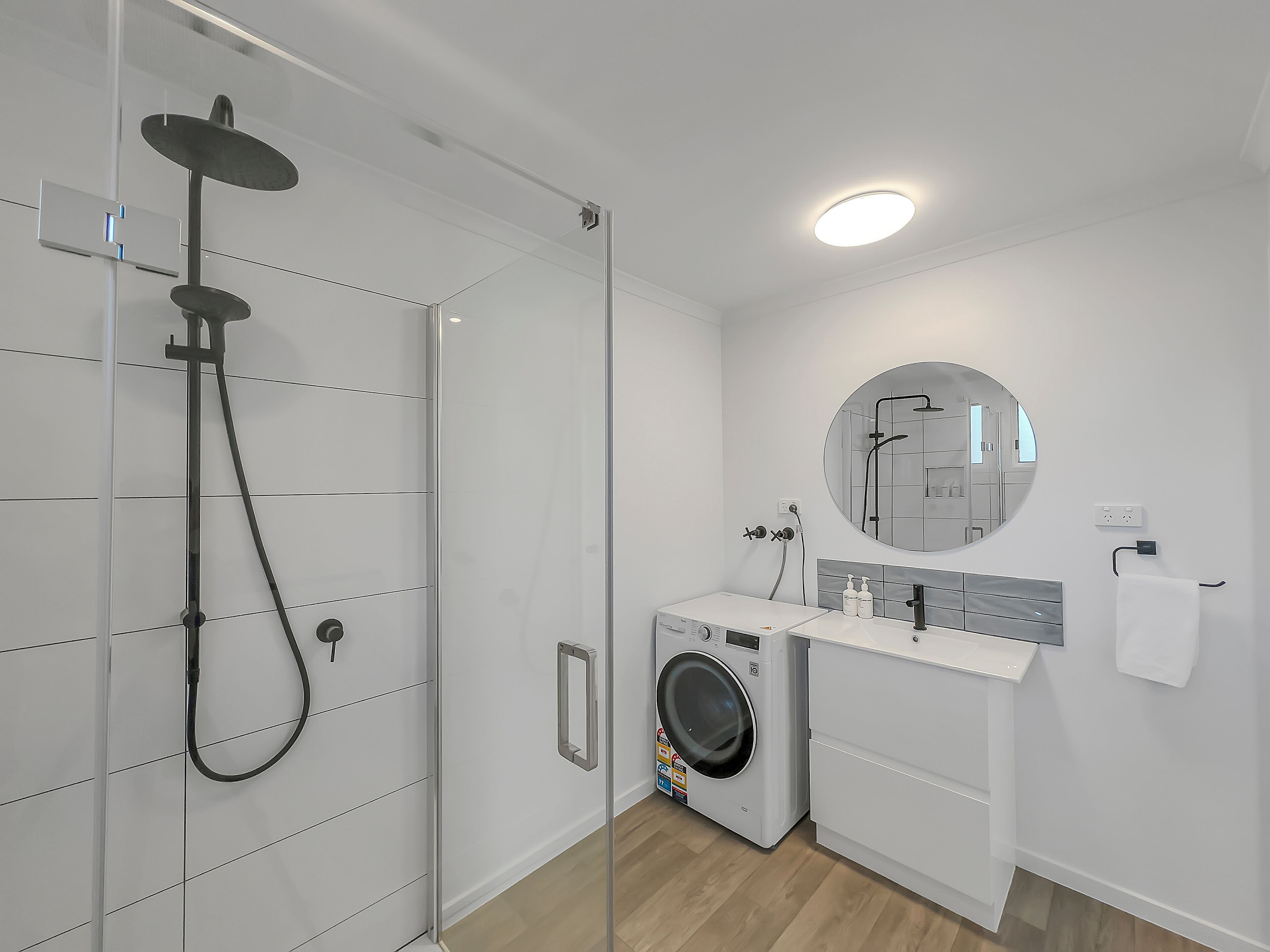 Een moderne badkamer met een glazen douchecabine, een ingebouwde wasmachine en een ronde spiegel boven de wastafel.