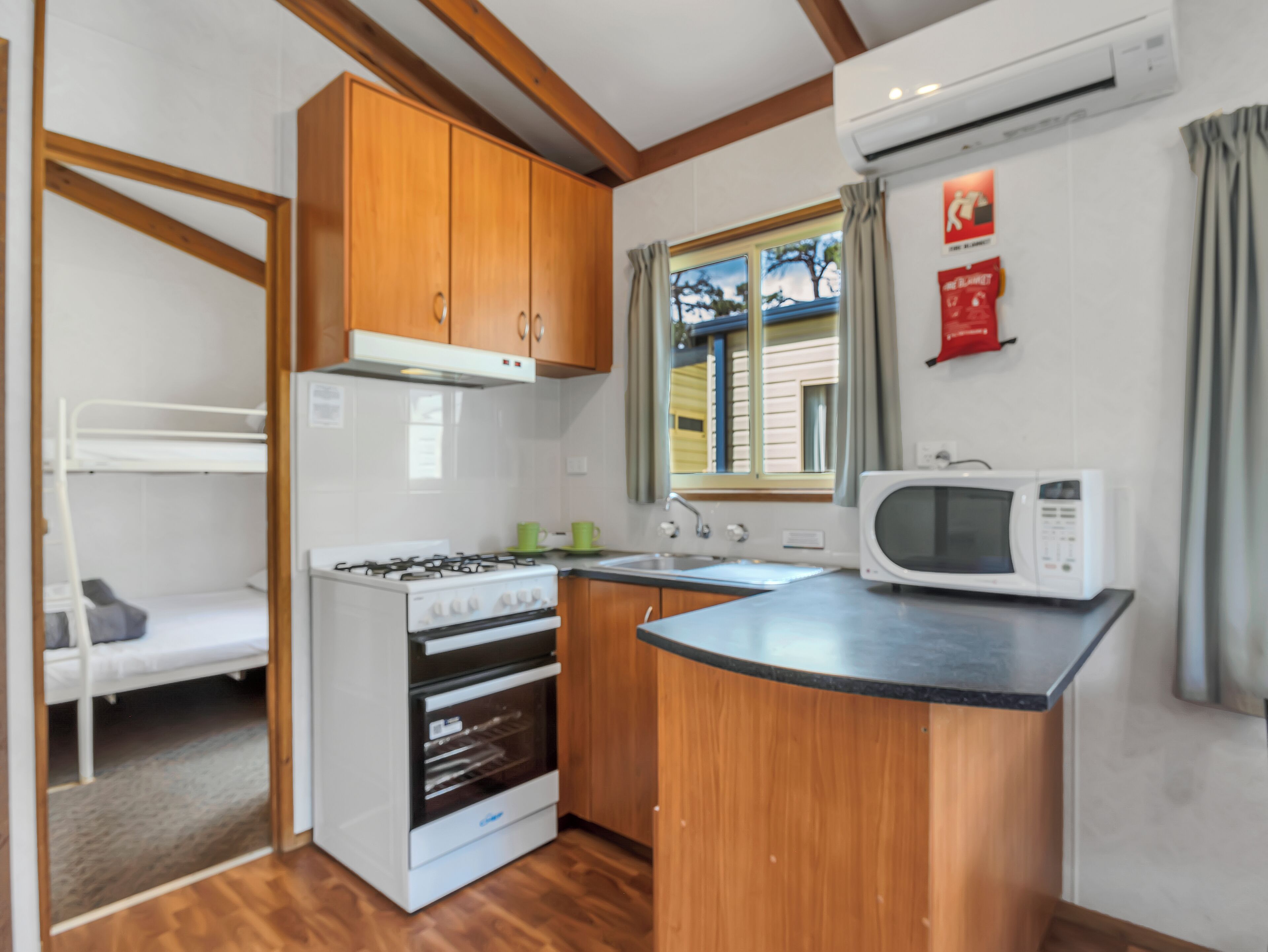 A compact kitchen with wooden cabinets, a gas stove, a microwave, and a window with curtains.