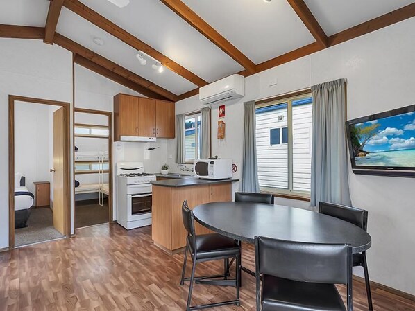 A compact kitchen with wooden cabinets, a stove, and a microwave. There is a dining table with chairs and a large window with curtains. - Ingenia Holidays Torquay (Torquay)