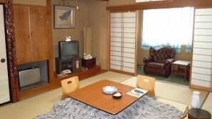 Ein traditioneller japanischer Raum mit Tatami-Bodenbelag, einem niedrigen Tisch mit Kissen, einem Fernseher und Schiebetüren.
