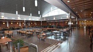 Uma ampla cafeteria interna com várias mesas e cadeiras, escada e teto alto com luminárias pendentes.