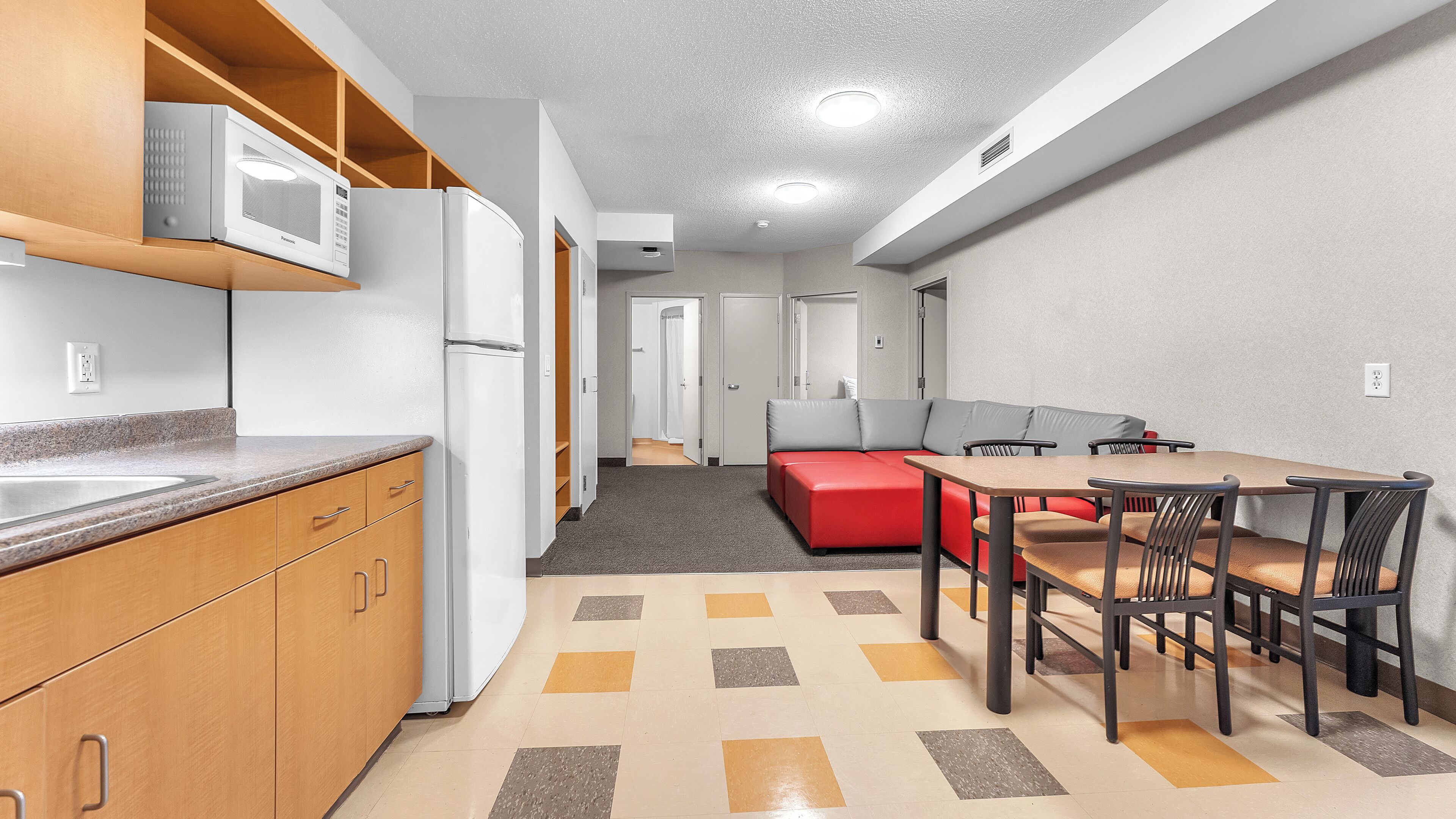 A compact kitchen with a microwave, refrigerator, and wooden cabinets. A dining area with a table and chairs is adjacent to a red sofa.