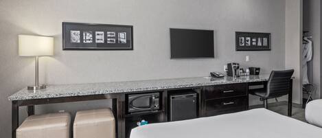 A hotel room with a bed, a desk with a lamp, a television, and a minibar.