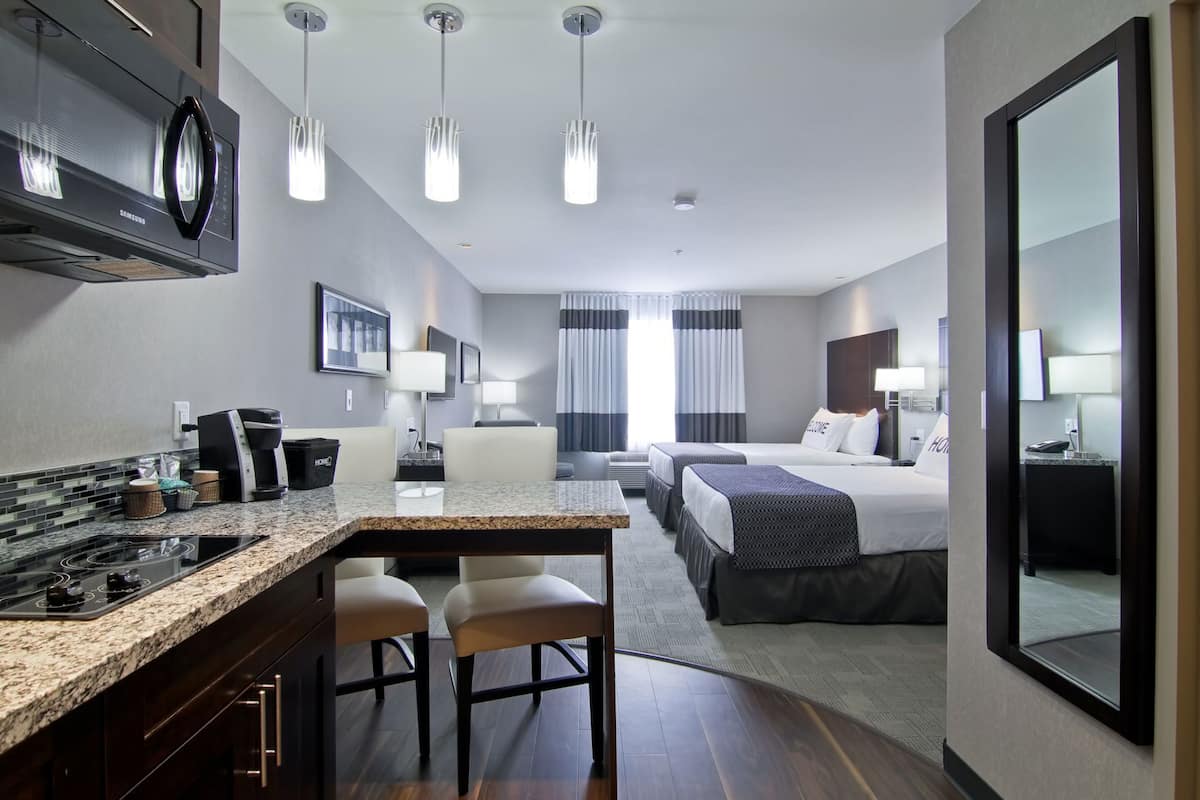 A hotel room with a kitchen area, a dining table, two beds, and a large mirror.