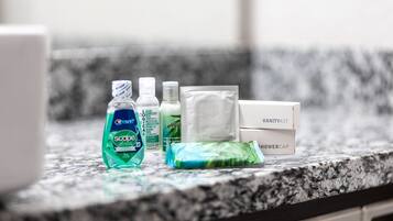 Una encimera de baño con varios artículos de aseo, incluyendo una botella de enjuague bucal Scope, un dispensador de gel antibacterial, un paquete de toallitas faciales, una botella de loción corporal y un gorro de ducha.