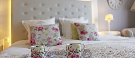 A neatly made bed with floral bedding, a tray with two cups and a glass, and a clock on the wall.