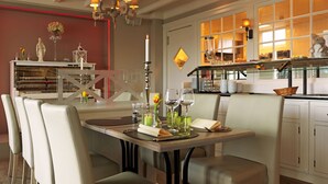 A dining area with a table set for four, chairs, a chandelier, and a kitchen in the background. - Ara Dune Hotel (De Panne)