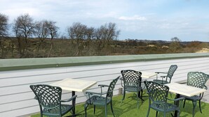 An outdoor patio with green metal chairs and tables, surrounded by a white fence and a view of trees and distant buildings. - Ara Dune Hotel (De Panne)