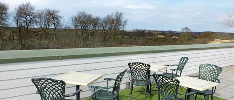 Un patio al aire libre con sillas y mesas de metal verde, rodeado por una cerca blanca y vistas a árboles y edificios distantes.