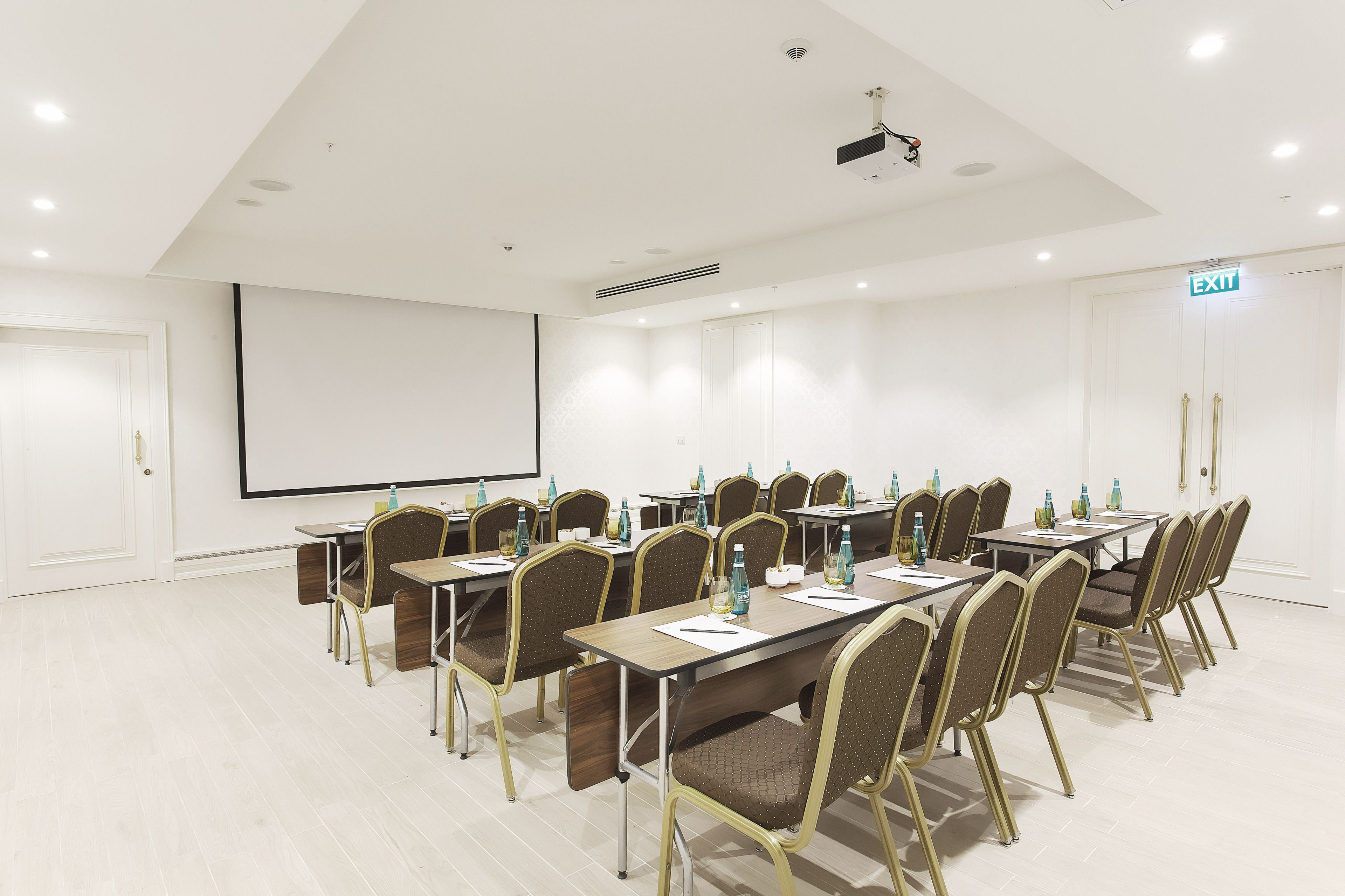A conference room with multiple tables and chairs, a projector screen, and an exit sign.