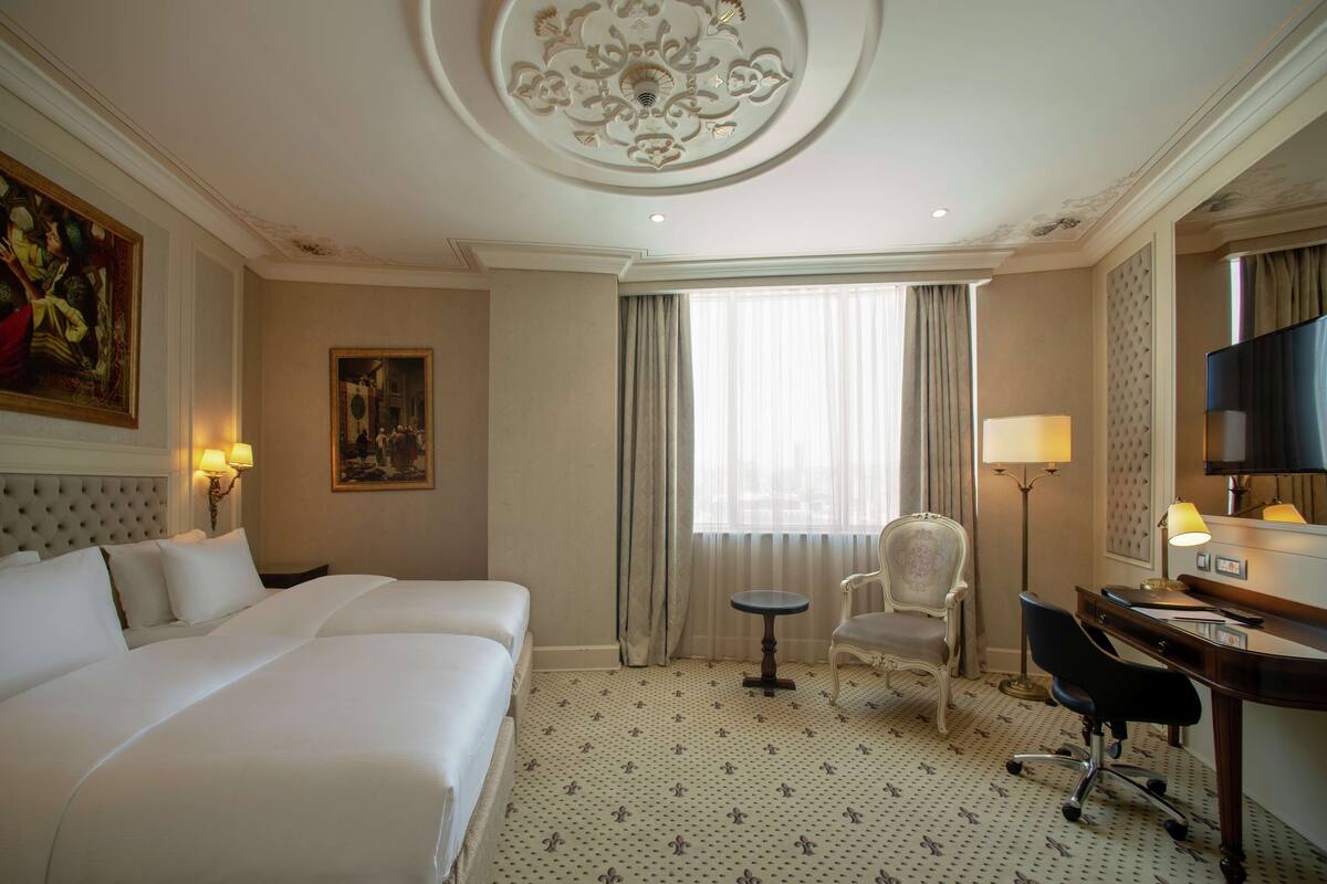 A hotel room with two beds, a desk, a chair, a television, and a large window with curtains.