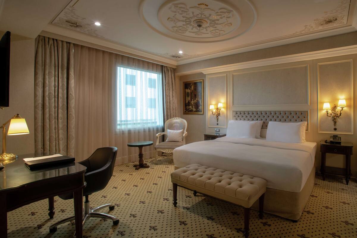 A hotel room with a large bed, a desk with a lamp, a chair, and a small table.