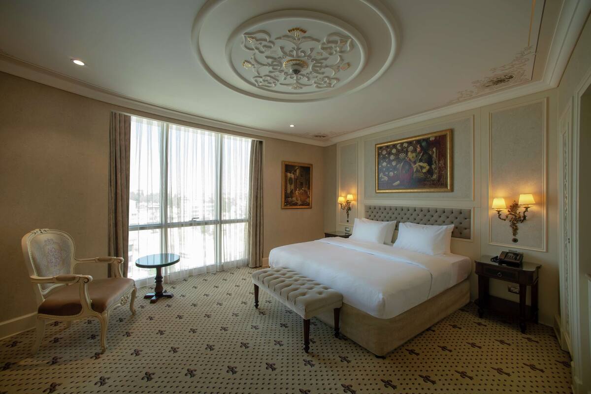A hotel room with a large bed, a chair, a small table, and a painting on the wall.
