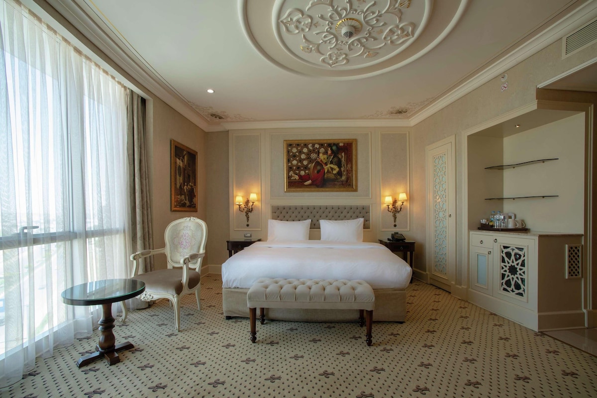 Un espacioso dormitorio con una cama grande, una zona de estar con una silla, una mesita pequeña y un techo decorativo.