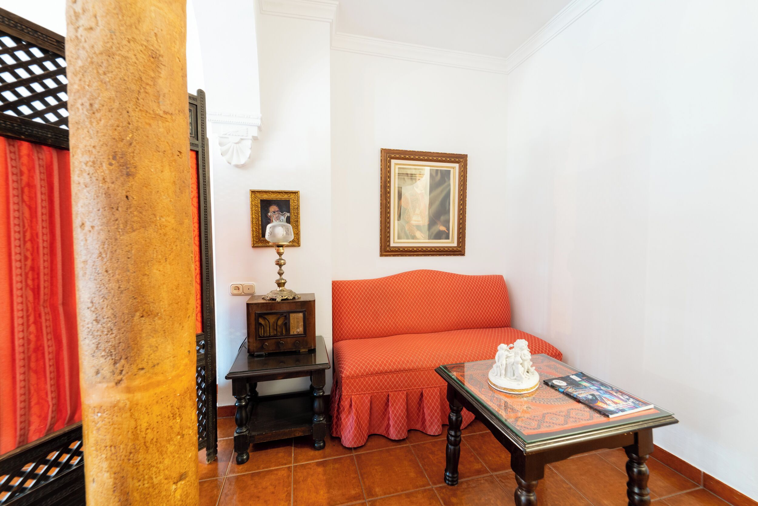 A room with a red sofa, a wooden table with a statue, a framed picture, and a lamp on a wooden cabinet.