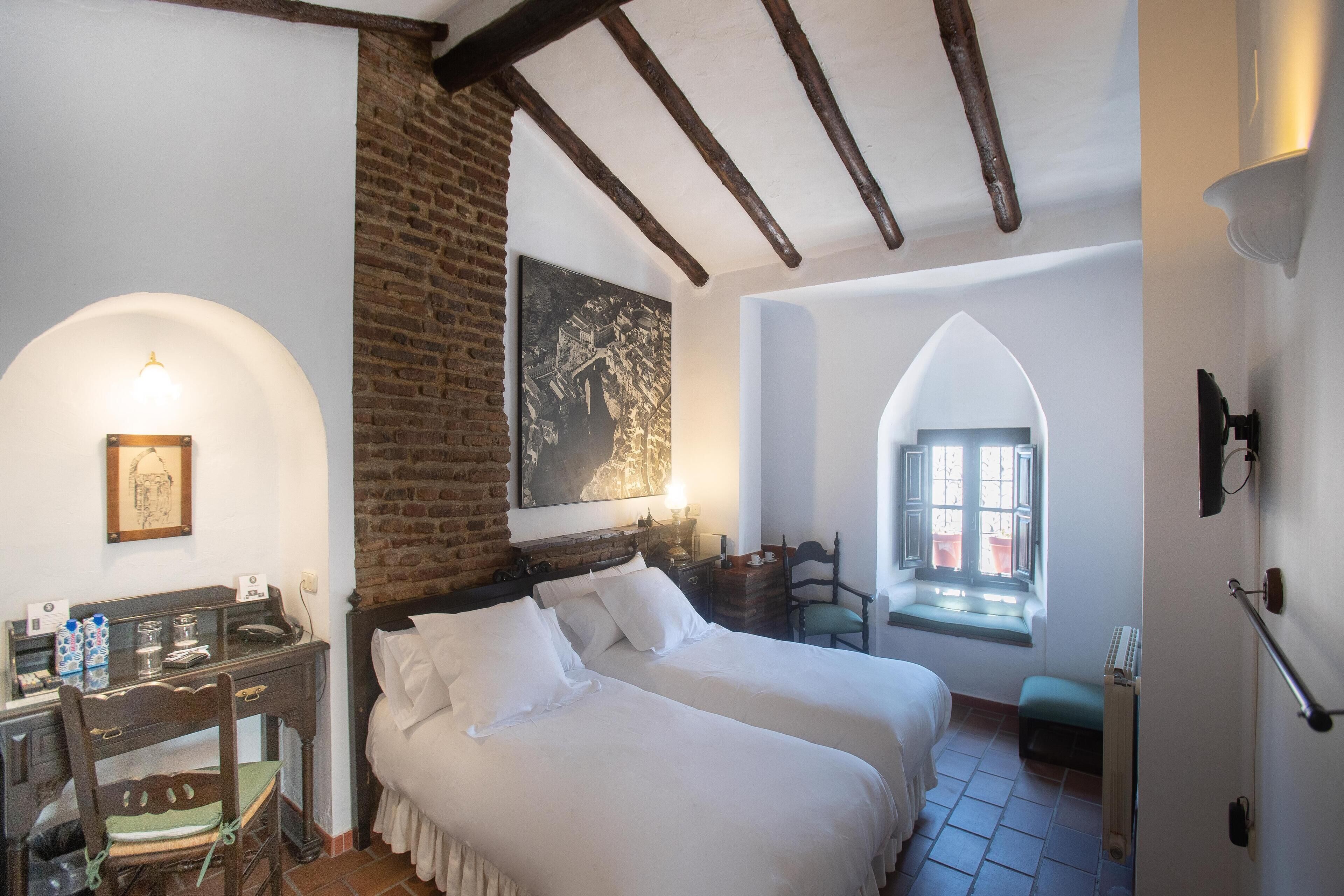 Una habitación con cama, escritorio, silla, ventana y pared de ladrillo.