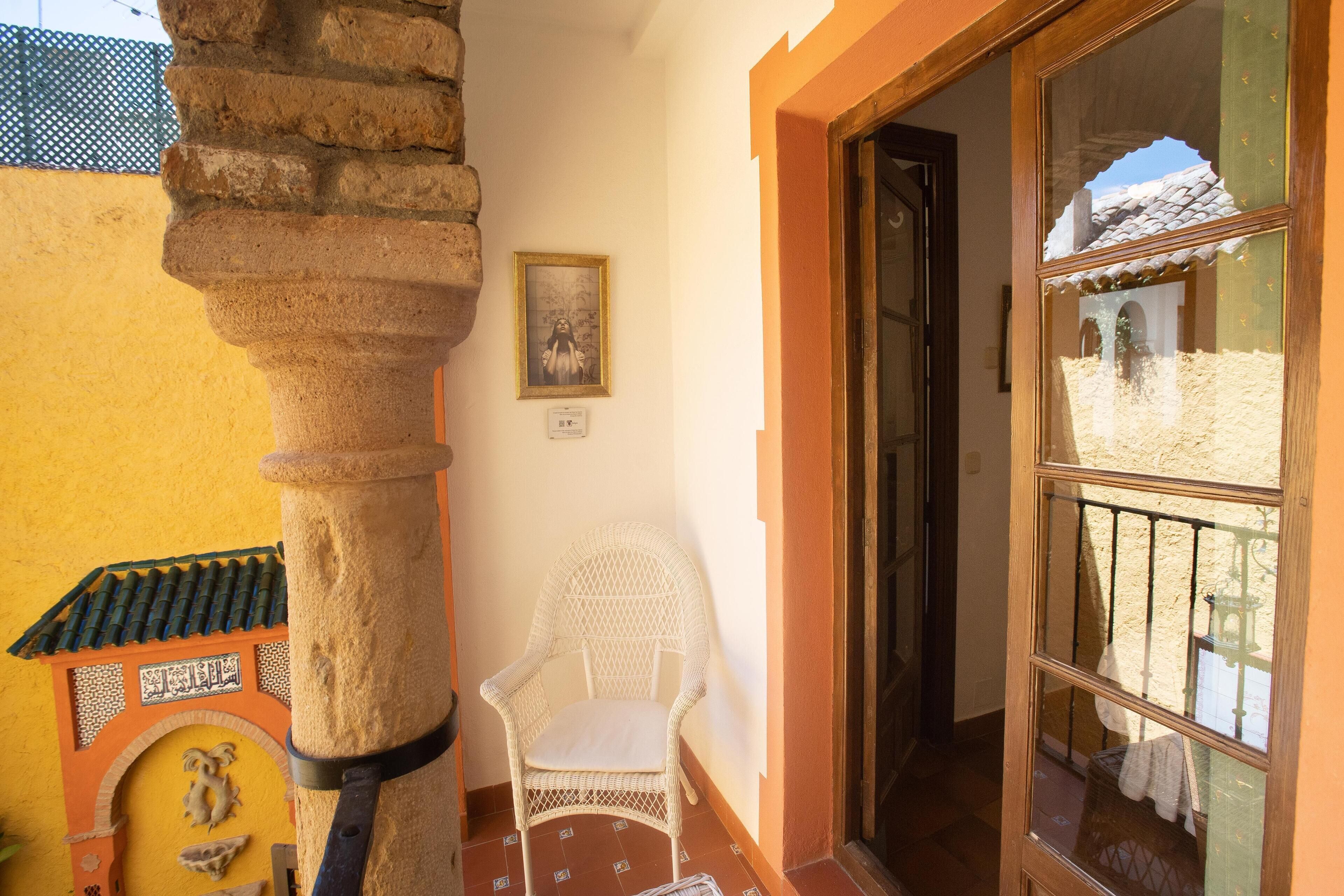 Une pièce avec un pilier en pierre, une chaise en osier blanche, un tableau encadré au mur et une fenêtre donnant sur une cour.