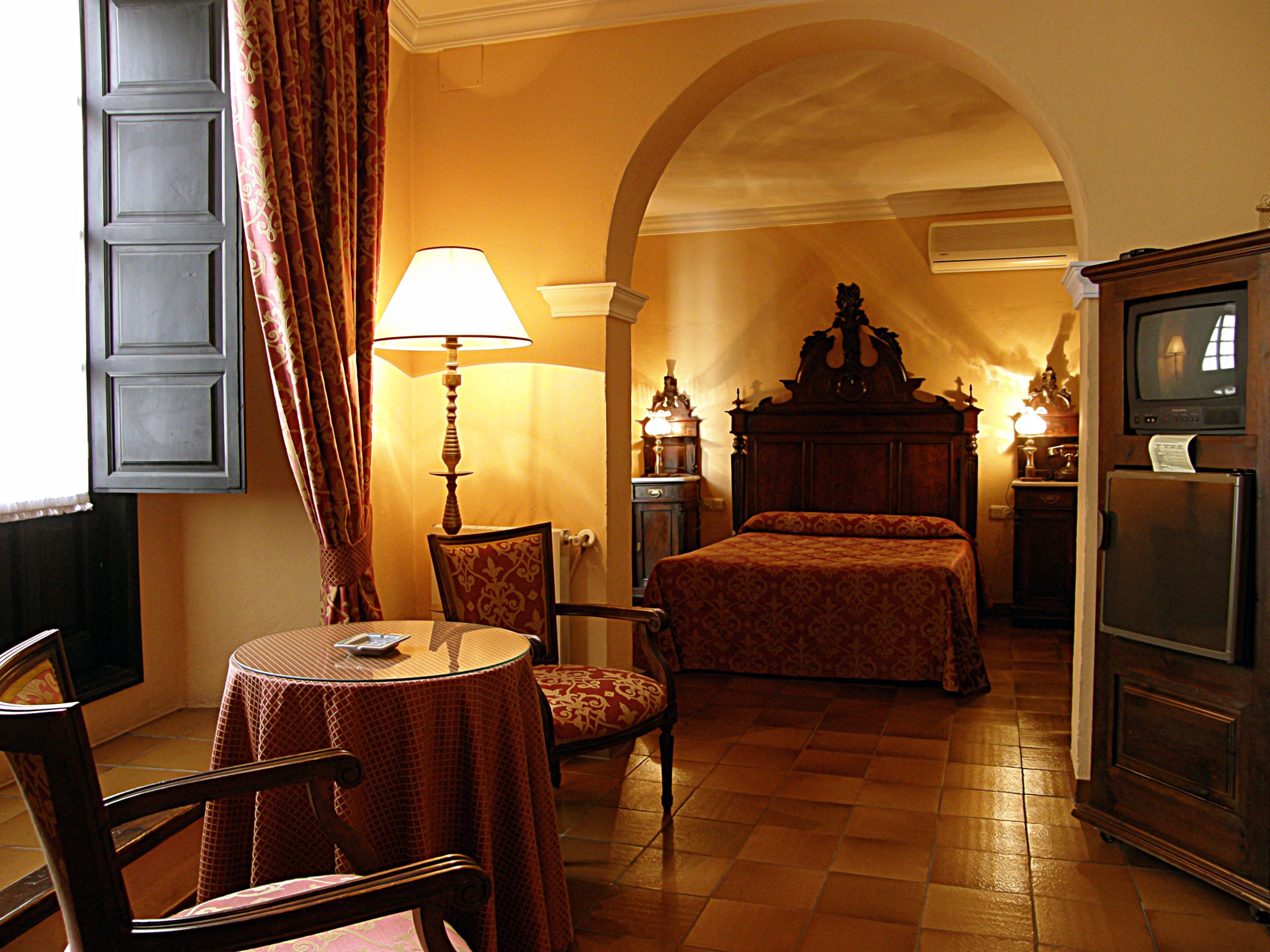 A hotel room with a bed, a table with chairs, a TV, and a lamp.