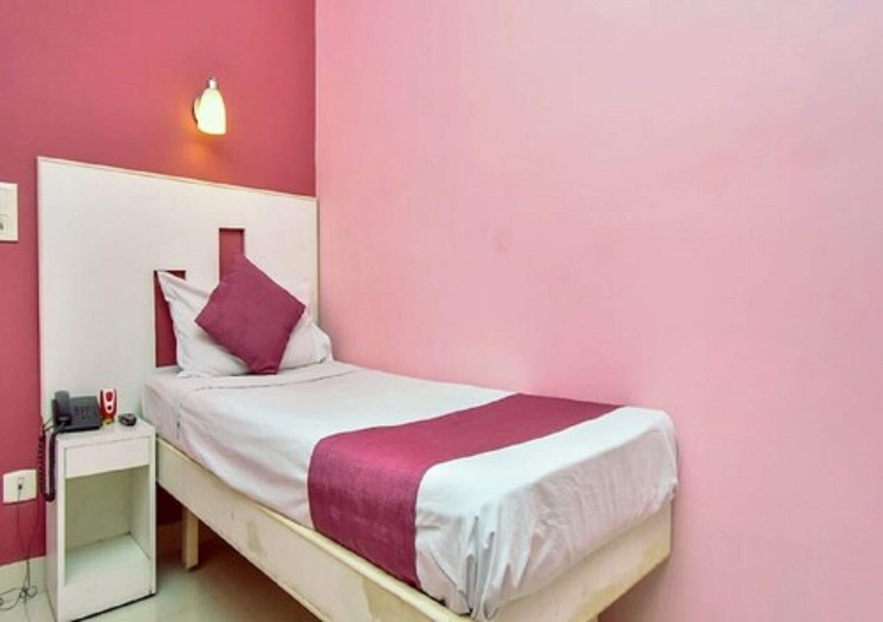 A single bed with a white and pink bedspread, a white nightstand with a lamp, and a pink wall.