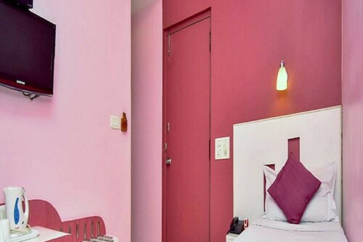 A pink-themed hotel room with a bed, a bedside table with a kettle, a TV mounted on the wall, and a door.