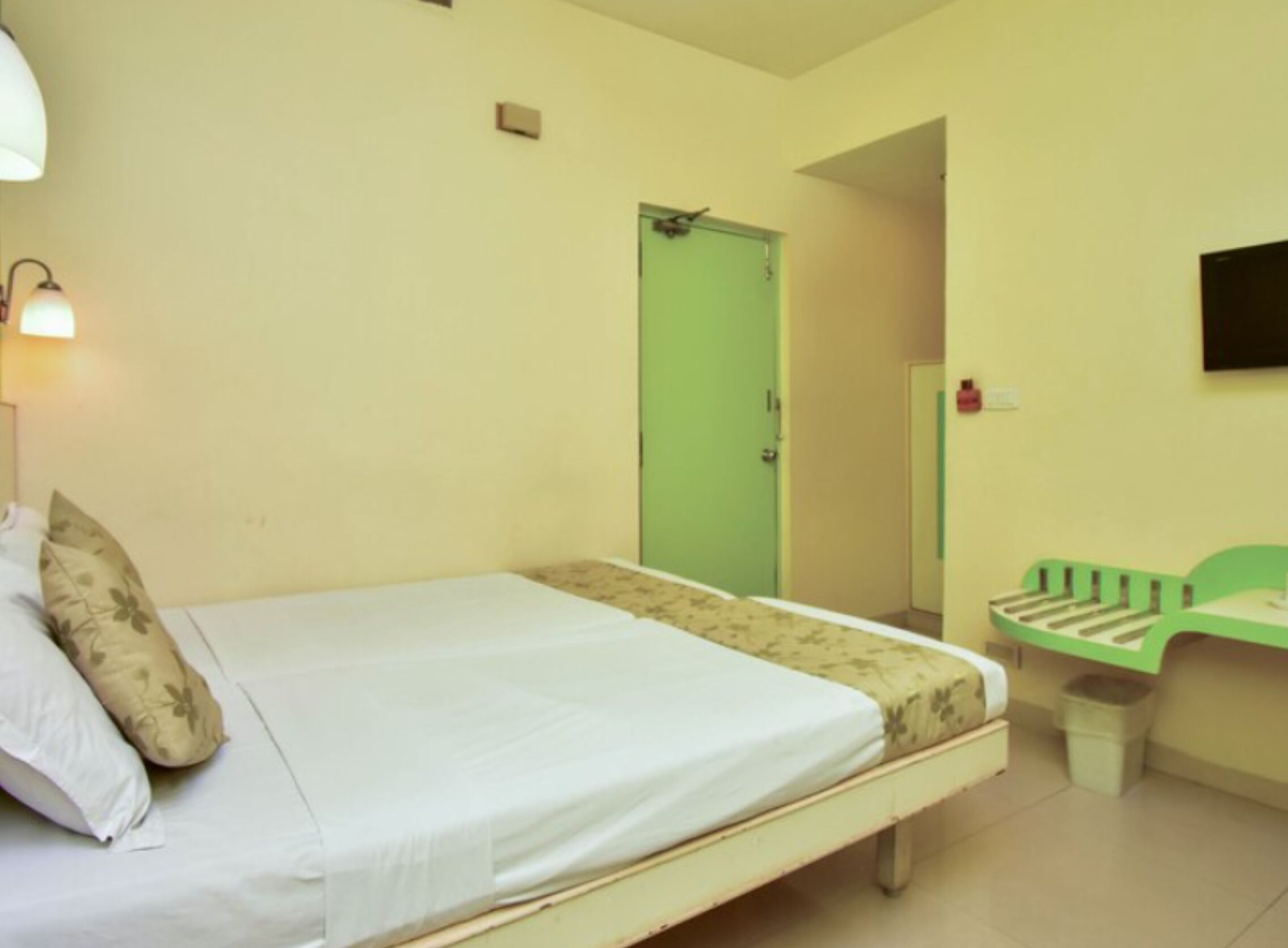 A single-bed room with a green door, a small desk with a chair, and a wall-mounted TV.