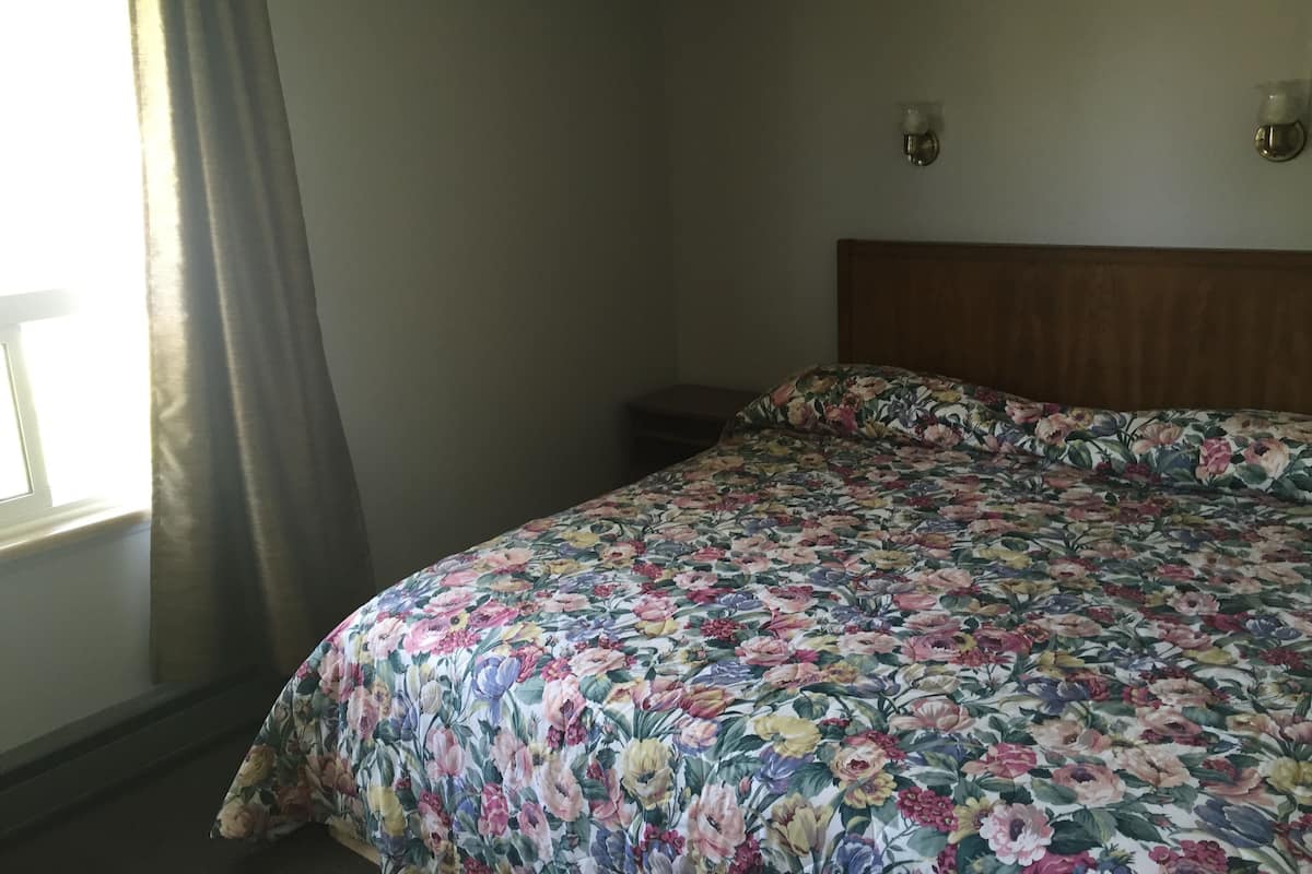 A bedroom with a floral bedspread, a wooden headboard, a bedside table, and a window with curtains.